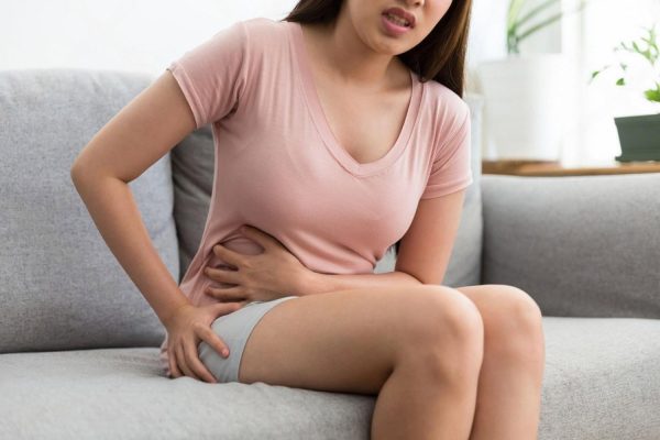 Young woman sitting on a sofa clutching her abdomen in pain, a warning sign of digestive disease